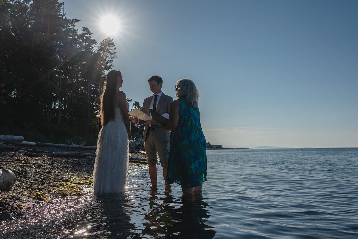 A couple stands ankle-deep in the ocean with their officiant as the sun flares over the shoreline